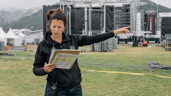Eventplaner mit Klemmbrett und Schichttabelle vor leerem Festivalgelände am Vortag